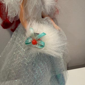 Puede incluir: Una mu&ntilde;eca con un vestido azul claro con una capa de purpurina plateada. La mu&ntilde;eca tiene el pelo rojo y un cuello blanco y esponjoso. Un peque&ntilde;o lazo verde azulado con una cuenta roja est&aacute; unido al cuello.