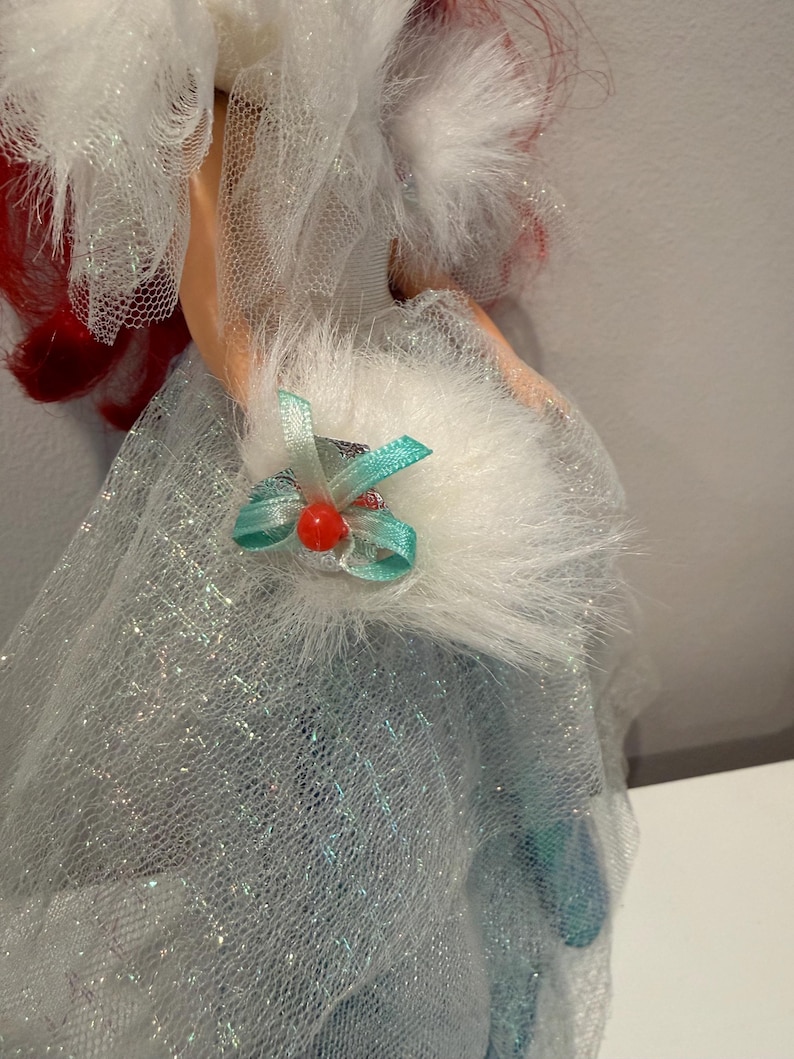Puede incluir: Una mu&ntilde;eca con un vestido azul claro con una capa de purpurina plateada. La mu&ntilde;eca tiene el pelo rojo y un cuello blanco y esponjoso. Un peque&ntilde;o lazo verde azulado con una cuenta roja est&aacute; unido al cuello.