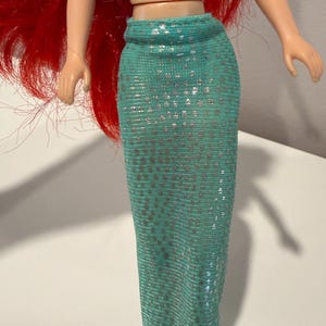 May include: A doll with red hair and a turquoise mermaid tail. The tail is covered in silver sequins. The doll's arms are outstretched, and the skin is a light peach color.