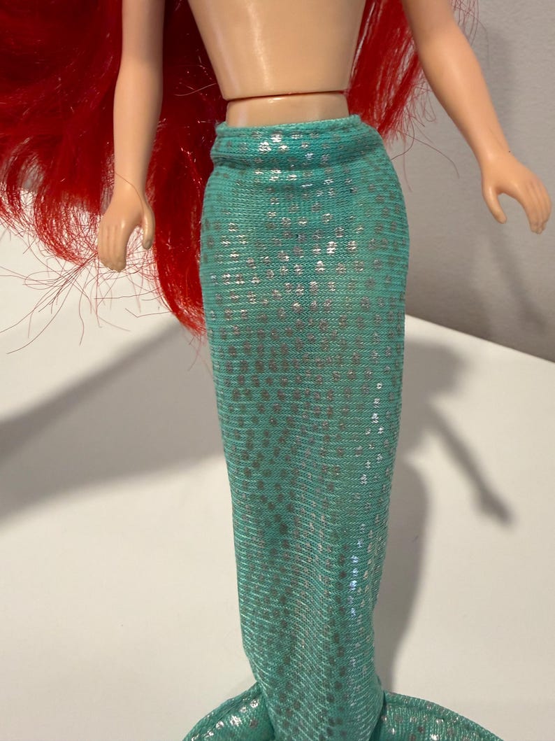 May include: A doll with red hair and a turquoise mermaid tail. The tail is covered in silver sequins. The doll's arms are outstretched, and the skin is a light peach color.