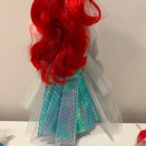 Puede incluir: Mu&ntilde;eca con cabello rojo vibrante y falda turquesa e iridiscente. El vestido de la mu&ntilde;eca tiene una capa blanca transparente, sobre un fondo blanco.