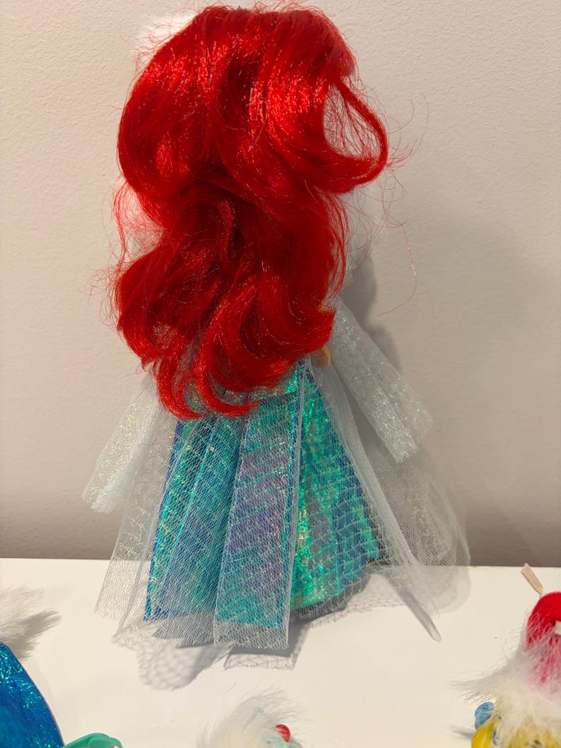 Puede incluir: Mu&ntilde;eca con cabello rojo vibrante y falda turquesa e iridiscente. El vestido de la mu&ntilde;eca tiene una capa blanca transparente, sobre un fondo blanco.