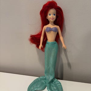 May include: A doll of a mermaid with red hair, blue eyes, and a purple seashell top. The mermaid has a teal sequined tail. The doll is standing on a white surface against a gray background.