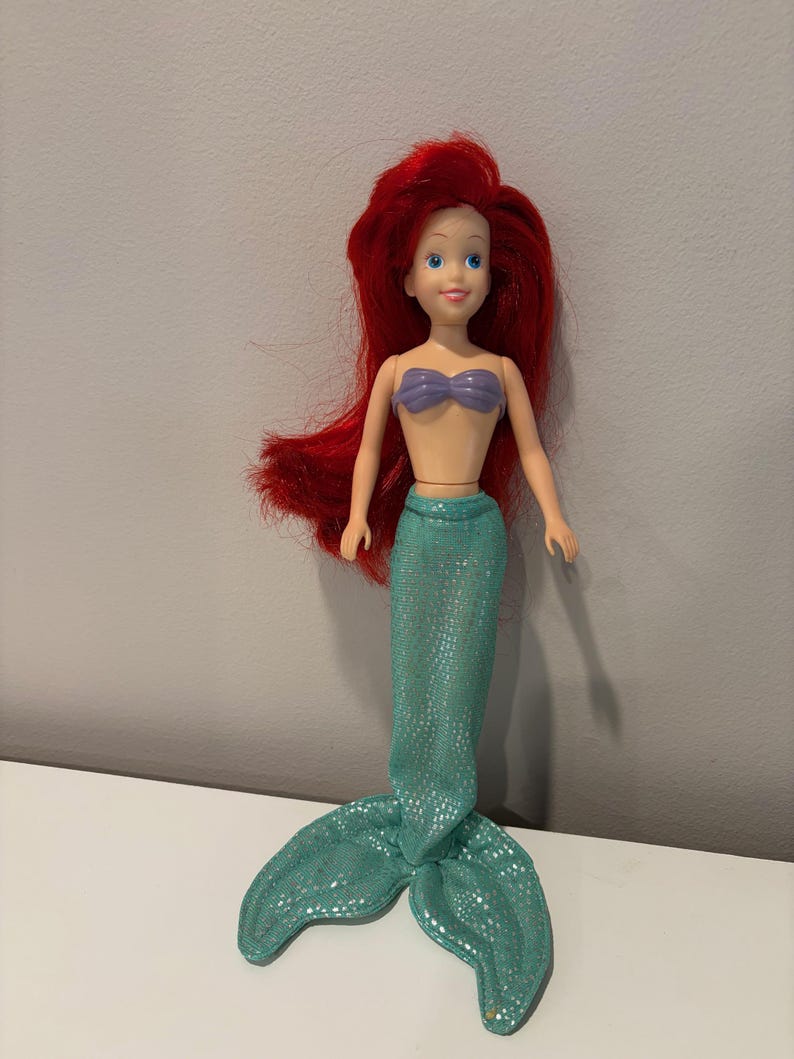 May include: A doll of a mermaid with red hair, blue eyes, and a purple seashell top. The mermaid has a teal sequined tail. The doll is standing on a white surface against a gray background.