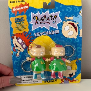 May include: Two Rugrats keychains on a blue and yellow Nickelodeon-branded card. The keychains feature Phil and Lil, each wearing a green dress and red shoes. The card reads "Rugrats KEYCHAINS" and "Ages 5 And Up."