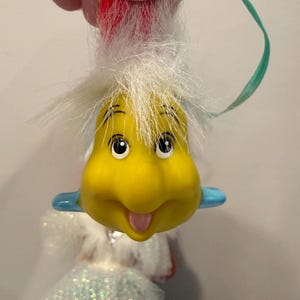 Puede incluir: Un adorno amarillo con forma de caricatura con una cara sonriente, ojos negros y una lengua rosa. Tiene pelo blanco y detalles azules. Se adjunta una cinta verde para colgarlo. El adorno es parte de una mu&ntilde;eca.
