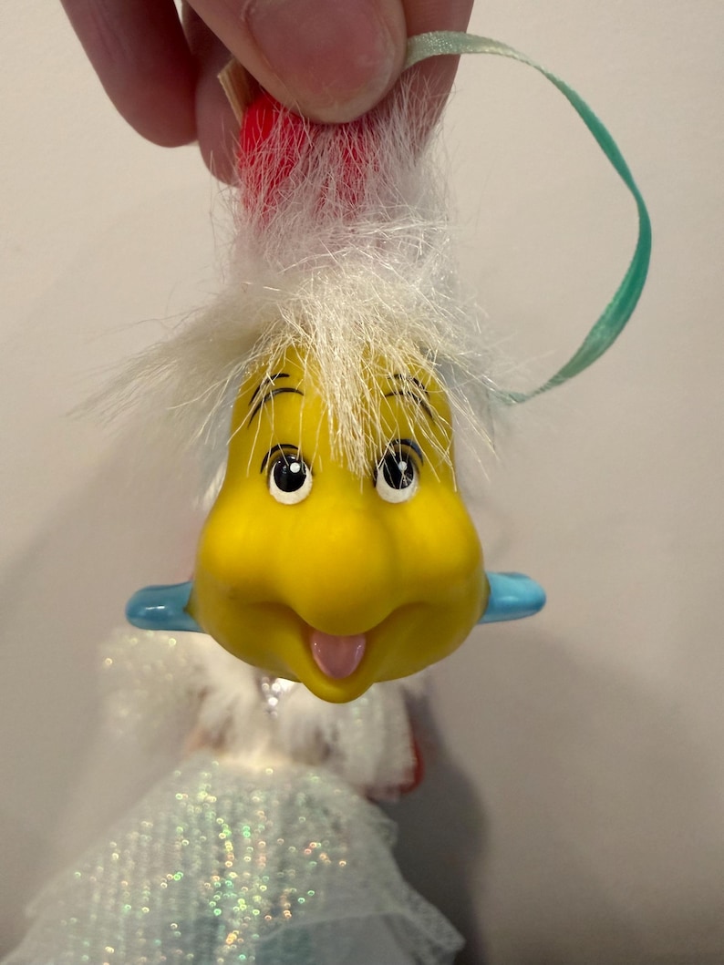 Puede incluir: Un adorno amarillo con forma de caricatura con una cara sonriente, ojos negros y una lengua rosa. Tiene pelo blanco y detalles azules. Se adjunta una cinta verde para colgarlo. El adorno es parte de una mu&ntilde;eca.