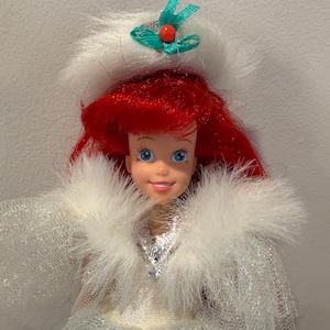 Puede incluir: Mu&ntilde;eca con cabello rojo y ojos azules, vestida con un atuendo ribeteado de piel blanca con un corpi&ntilde;o plateado y un sombrero blanco con un lazo verde y una cuenta roja. La mu&ntilde;eca es un personaje de una pel&iacute;cula de Disney.