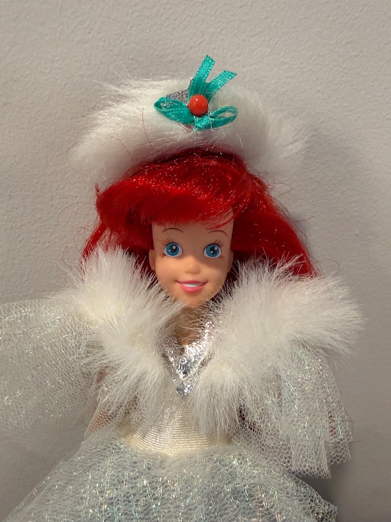 Puede incluir: Mu&ntilde;eca con cabello rojo y ojos azules, vestida con un atuendo ribeteado de piel blanca con un corpi&ntilde;o plateado y un sombrero blanco con un lazo verde y una cuenta roja. La mu&ntilde;eca es un personaje de una pel&iacute;cula de Disney.