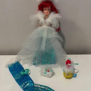Puede incluir: Mu&ntilde;eca con cabello rojo, vestida con un abrigo ribeteado de piel blanca y un vestido azul y blanco. Accesorios: cola de sirena, sombrero peque&ntilde;o y figura de pez amarillo. La mu&ntilde;eca mide unos 29 cm de alto.
