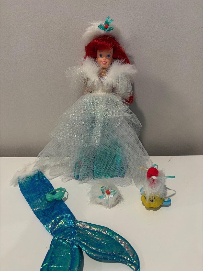 Puede incluir: Mu&ntilde;eca con cabello rojo, vestida con un abrigo ribeteado de piel blanca y un vestido azul y blanco. Accesorios: cola de sirena, sombrero peque&ntilde;o y figura de pez amarillo. La mu&ntilde;eca mide unos 29 cm de alto.