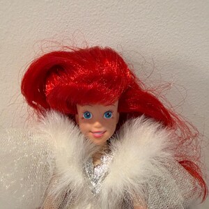 Puede incluir: Mu&ntilde;eca con cabello rojo brillante y ojos azules. La mu&ntilde;eca lleva un vestido plateado y un chal blanco y esponjoso de tul. La piel de la mu&ntilde;eca es de color melocot&oacute;n claro. La mu&ntilde;eca es un personaje popular de una pel&iacute;cula infantil.