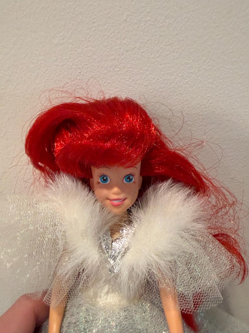 Puede incluir: Mu&ntilde;eca con cabello rojo brillante y ojos azules. La mu&ntilde;eca lleva un vestido plateado y un chal blanco y esponjoso de tul. La piel de la mu&ntilde;eca es de color melocot&oacute;n claro. La mu&ntilde;eca es un personaje popular de una pel&iacute;cula infantil.