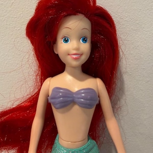 May include: A plastic doll of a mermaid with bright red hair, blue eyes, and a purple seashell top. The doll has a light skin tone and a teal green skirt. The doll is a popular character from a children's animated movie.