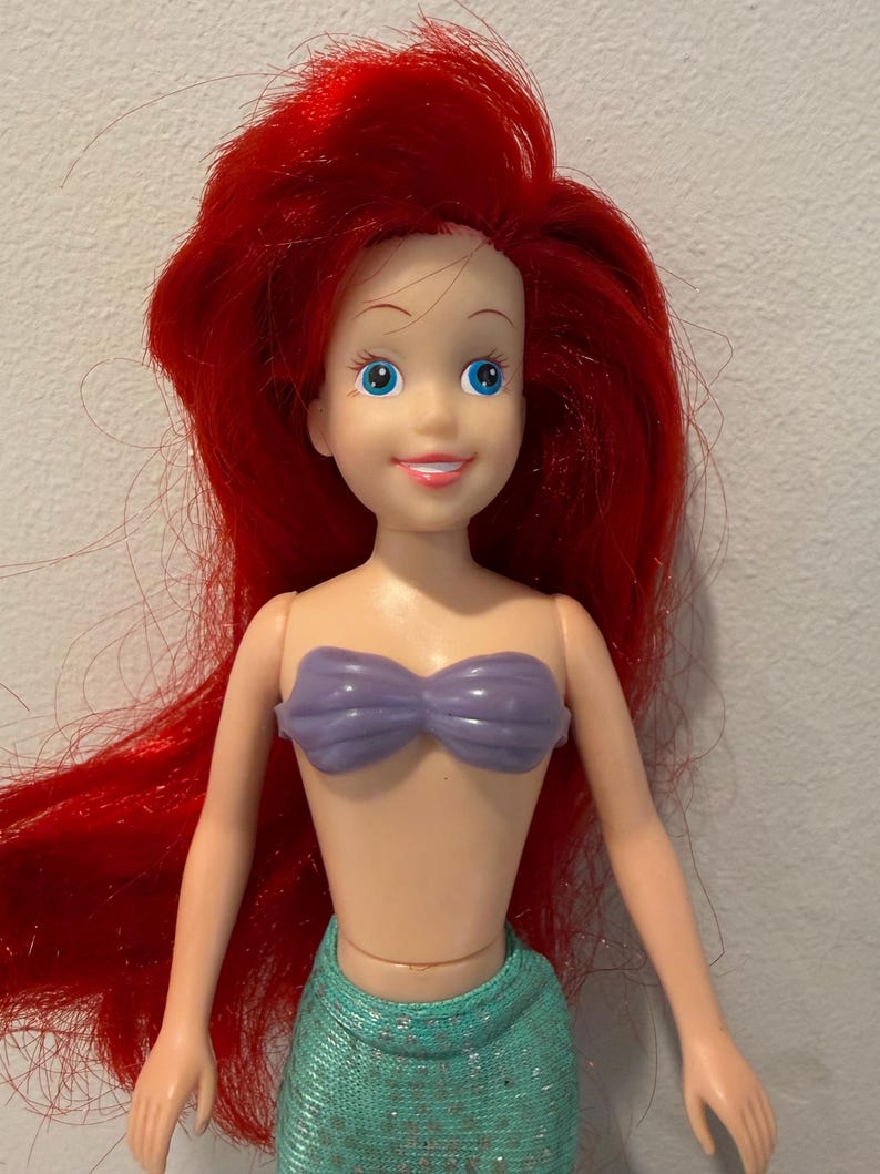 May include: A plastic doll of a mermaid with bright red hair, blue eyes, and a purple seashell top. The doll has a light skin tone and a teal green skirt. The doll is a popular character from a children's animated movie.
