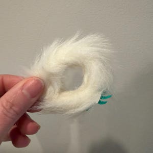 Puede incluir: Un objeto en forma de anillo, de color blanco y esponjoso, con un agujero en el centro. Tiene una textura suave y peluda. Se ven peque&ntilde;os detalles turquesas en un lado. El objeto est&aacute; sostenido por una mano.
