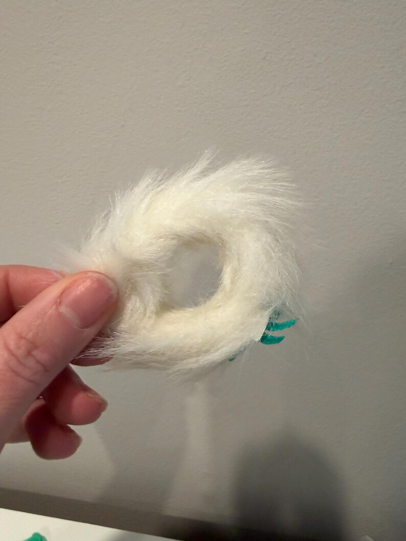 Puede incluir: Un objeto en forma de anillo, de color blanco y esponjoso, con un agujero en el centro. Tiene una textura suave y peluda. Se ven peque&ntilde;os detalles turquesas en un lado. El objeto est&aacute; sostenido por una mano.