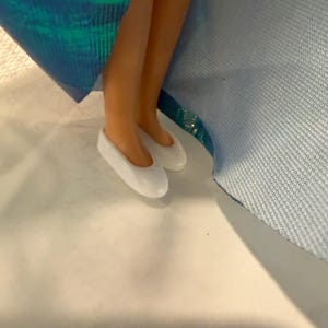 Puede incluir: Piernas y pies de mu&ntilde;eca con bailarinas blancas. La mu&ntilde;eca lleva un vestido iridiscente azul y verde. La imagen est&aacute; tomada desde un &aacute;ngulo bajo.