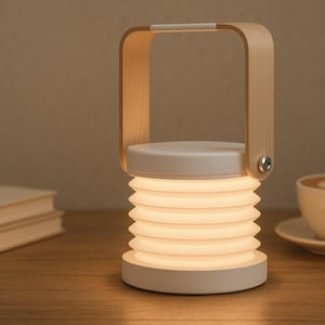 May include: A white and wood-tone LED lantern with a collapsible, accordion-style shade. The lantern has a wooden handle and sits on a wooden surface. A cup of coffee and books are in the background.