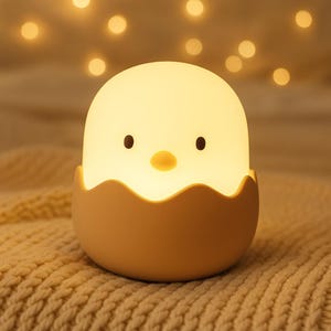 May include: A soft yellow night light shaped like a chick hatching from an egg. The chick has black eyes and a yellow beak. The eggshell base is a light tan color. The light is glowing, and the background is blurred with soft, warm lights.