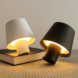 Peut inclure: Deux lampes de table modernes, une blanche et une noire, avec un abat-jour conique et une base cylindrique. Les lampes sont allumées, dégageant une lueur chaleureuse. Elles reposent sur un livre fermé, suggérant un cadre de chevet ou de bureau.