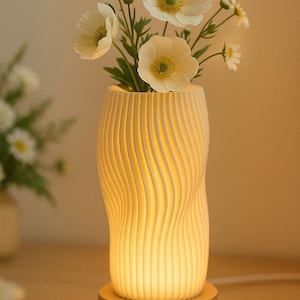 Pode incluir: Um vaso de cerâmica branco com um design ondulado e texturizado, iluminado por dentro, assenta numa base de madeira. Contém flores brancas com centros amarelos e caules verdes, criando um brilho suave. A lâmpada é um elemento decorativo.