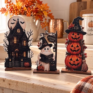 May include: Three Halloween-themed wooden tabletop decorations on rectangular bases. The left piece is a dark haunted house with a full moon and bats. The centre piece is a ghost wearing a witch hat, holding a book labelled Spells, with a sign reading Witching Hour. The right piece is a stack of three orange jack-o'-lanterns wearing a witch hat and holding a broom.