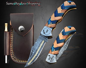 Op de afbeelding: Een vouwmes met een houten handvat met blauwe en bruine accenten en een patroon op het lemmet. Het mes wordt getoond met een bruine leren schede en een vuurstarter. Het lemmet heeft de initialen "A&B" en "Happy Birthday Anthony".