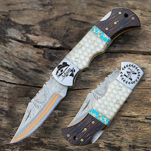 May include: Two folding pocket knives with Damascus steel blades featuring orange accents. The handles have dark wood ends, a central white honeycomb pattern section, and turquoise spacers. One blade is engraved with a fish and the name Adam.