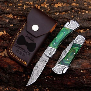 May include: A brown leather knife case with the name "Justin", a bow tie graphic, and the text "Groomsman 02-28-23" is next to a folding knife with a green handle and a decorative silver bolster. The knife is open and closed.