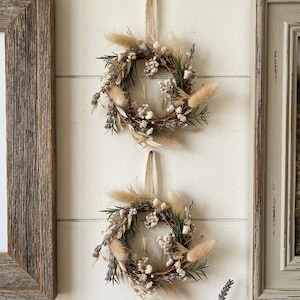 May include: Two small, round wreaths made of dried flowers and greenery hang on a white wall. The wreaths feature beige pampas grass, white flowers, and lavender accents. They are suspended by beige ribbons. The wall is decorated with framed pictures.