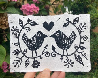 Op de afbeelding: Zwart-wit linoleumsnede met twee vogels die tegenover elkaar staan onder een hart. De vogels zijn omgeven door een bloemen- en bladerontwerp. De prent staat op getextureerd wit papier.