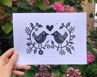 Op de afbeelding: Zwart-wit linoleumsnede met twee gestileerde vogels die tegenover elkaar staan, met een hart erboven. De vogels zijn omgeven door bloemen en bladeren. De tekst "Lovebirds - Black" staat onder de afbeelding.
