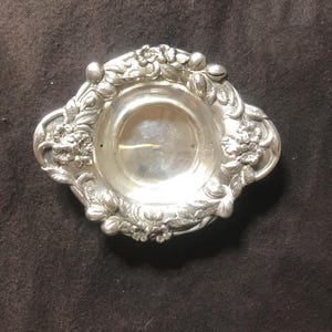 Op de afbeelding: Een kleine, zilverkleurige schaal met een bloemmotief rond de rand. De schaal heeft een ondiep, cirkelvormig midden en een sierlijke, verhoogde rand met gedetailleerde bloemen en bladeren. De algemene vorm is ovaal en de schaal lijkt van metaal te zijn.