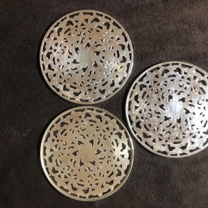 May include: Three silver-coloured, circular trivets with intricate, openwork designs. The trivets are arranged on a dark brown surface. Each trivet features a repeating pattern of floral and geometric shapes, creating a decorative and functional item.