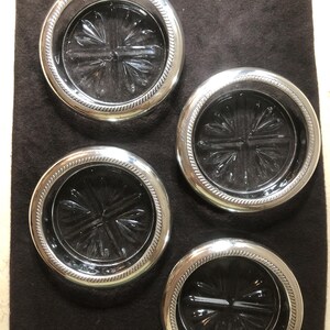 Op de afbeelding: Vier glazen onderzetters met zilveren randen en een stervormig patroon. De onderzetters hebben een decoratieve, touwachtige rand. Ze liggen op een donkerbruine achtergrond. Deze onderzetters beschermen oppervlakken tegen kringen.