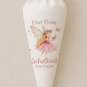 Peut inclure: Un sac cadeau en forme de cône en tissu crème, noué au sommet par un ruban assorti. L'avant présente une illustration imprimée d'une fée aux cheveux blonds avec un tutu rose, entourée de papillons, avec le texte Dein Name, Schulkind, Dein Datum.