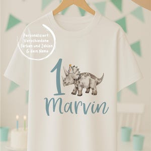 Könnte beinhalten: Ein weißes Kurzarm-T-Shirt mit einer Grafik eines grauen Triceratops mit Partyhut neben einer großen blauen Eins und dem Namen Marvin in blauer Schreibschrift. Der Text auf dem T-Shirt lautet: Personalisiert: Verschiedene Farben und Zahlen & dein Name.