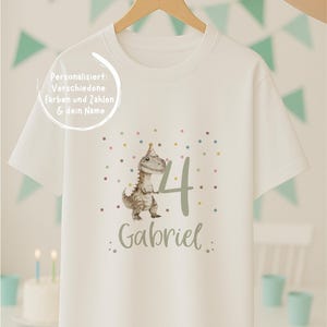 Peut inclure: Un t-shirt blanc à manches courtes orné d'un dinosaure portant un chapeau de fête à côté du chiffre 4 et du prénom Gabriel. Le motif inclut des points de confettis colorés. Le texte sur le t-shirt indique : Personalisiert: Verschiedene Farben und Zahlen & dein Name.