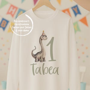 Könnte beinhalten: Ein langärmeliges cremefarbenes Shirt mit einer Grafik eines grauen Dinosauriers mit buntem Partyhut, der Zahl 1 in Salbeigrün und dem Namen Tabea in salbeigrüner Schreibschrift. Der Text auf dem Shirt lautet: Personalisiert: Verschiedene Farben und Zahlen & dein Name.