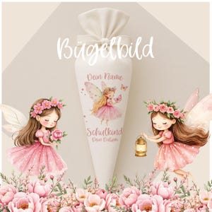 Op de afbeelding: Een witte stoffen kegel met een strik aan de bovenkant, voorzien van illustraties van twee feeën in roze jurken met bloemenkransen. De kegel toont de tekst Bügelbild, Dein Name, Schulkind en Dein Datum. Roze bloemen staan onderaan.