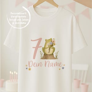 Könnte beinhalten: Weißes T-Shirt mit einem skurrilen Design mit einem grünen Frosch, der Blumen hält, der Zahl 7 in Rosa und den Worten "Dein Name". Das Shirt wird von einem Holzbügel gehalten.
