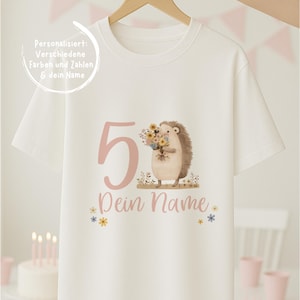 Puede incluir: Camiseta blanca con el número 5 rosa y un erizo de dibujos animados sosteniendo flores. El texto "Dein Name" está debajo del número, con pequeños gráficos de flores. La camiseta está colgada en una percha de madera.