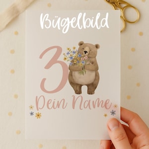 Könnte beinhalten: Eine Grußkarte mit dem deutschen Text "Bügelbild" und "3 Dein Name". Die Karte zeigt einen braunen Bären, der Blumen hält. Eine goldene Schere und ein Band sind im Hintergrund.