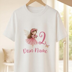 Op de afbeelding: Een wit T-shirt met korte mouwen en een afbeelding van een fee met bruin haar in een roze jurk met een bloemenkrans. Naast de fee staan een groot roze cijfer 2, een kleine vlinder en de roze tekst Dein Name onder de afbeelding.