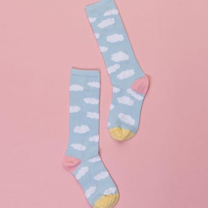 May include: Two light blue socks with a white cloud pattern. The socks have pink heels and yellow toes, set against a pink background. The socks are knee-high.