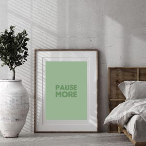Op de afbeelding: Een ingelijste print met de woorden "PAUSE MORE" in groene letters op een groene achtergrond. De lijst is bruin. Een witte vaas met een plant en een bed staan op de achtergrond.
