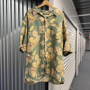 May include: A short-sleeved button-down shirt in a muted olive green featuring an all-over print of vintage-style travel labels and luggage tags. The pattern includes rectangular and circular graphics with text reading Shepheards Hotel and Cairo.