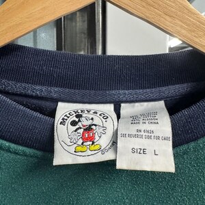 Puede incluir: Primer plano de un cuello de sudadera verde y azul marino con una etiqueta de Mickey & Co. La etiqueta presenta una imagen de dibujos animados de Mickey Mouse. Una segunda etiqueta indica la talla L.