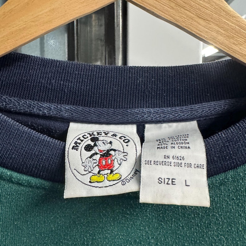 Puede incluir: Primer plano de un cuello de sudadera verde y azul marino con una etiqueta de Mickey & Co. La etiqueta presenta una imagen de dibujos animados de Mickey Mouse. Una segunda etiqueta indica la talla L.
